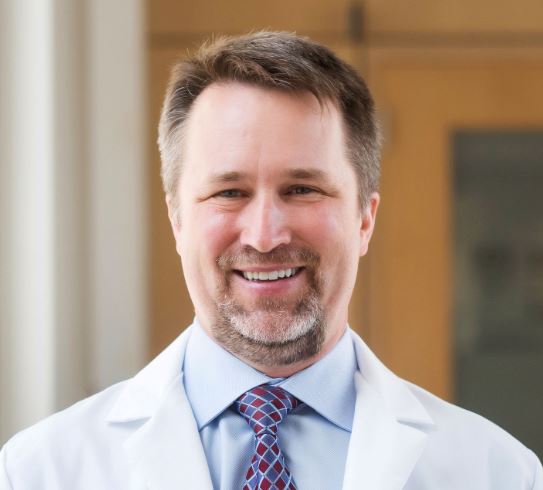 A man in a lab coat and tie poses for a headshot. 