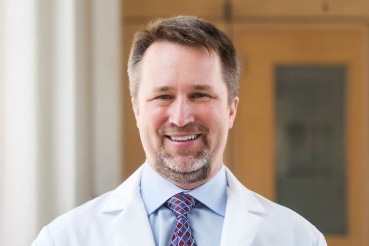 A man in a lab coat and tie poses for a headshot. 