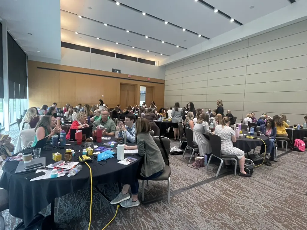 A large conference room full of people sitting at round tables is shown.