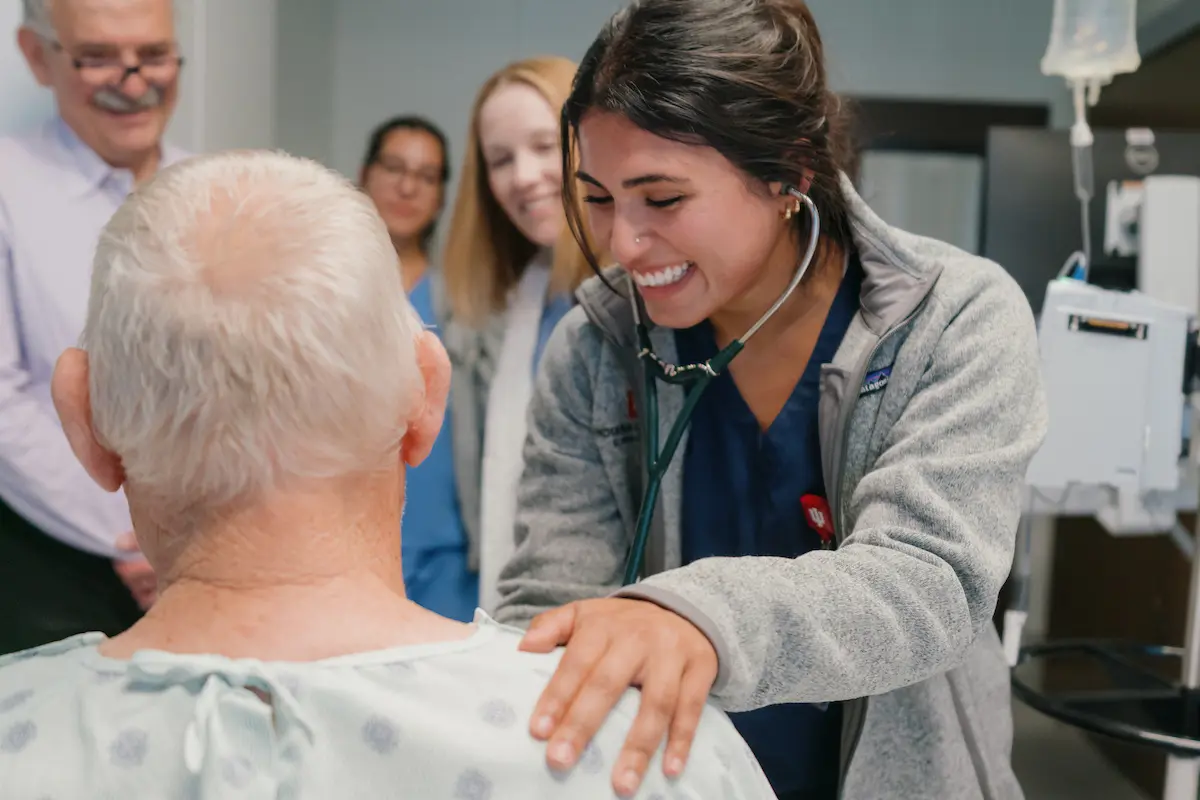 A medical student holds her hand on a patient's shoulder as she listens to his chest with a stethoscope. Two physicians behind her look on.