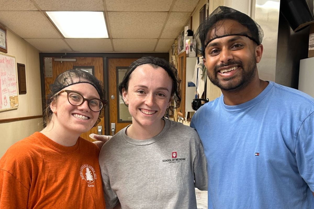 Three IU School of Medicine students smile as they wear gloves and hairnets, preparing to volunteer at St. Joseph's Soup Kitchen.