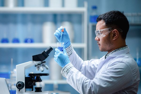 man in white coat looking at test tube in lab with microscope in front of him