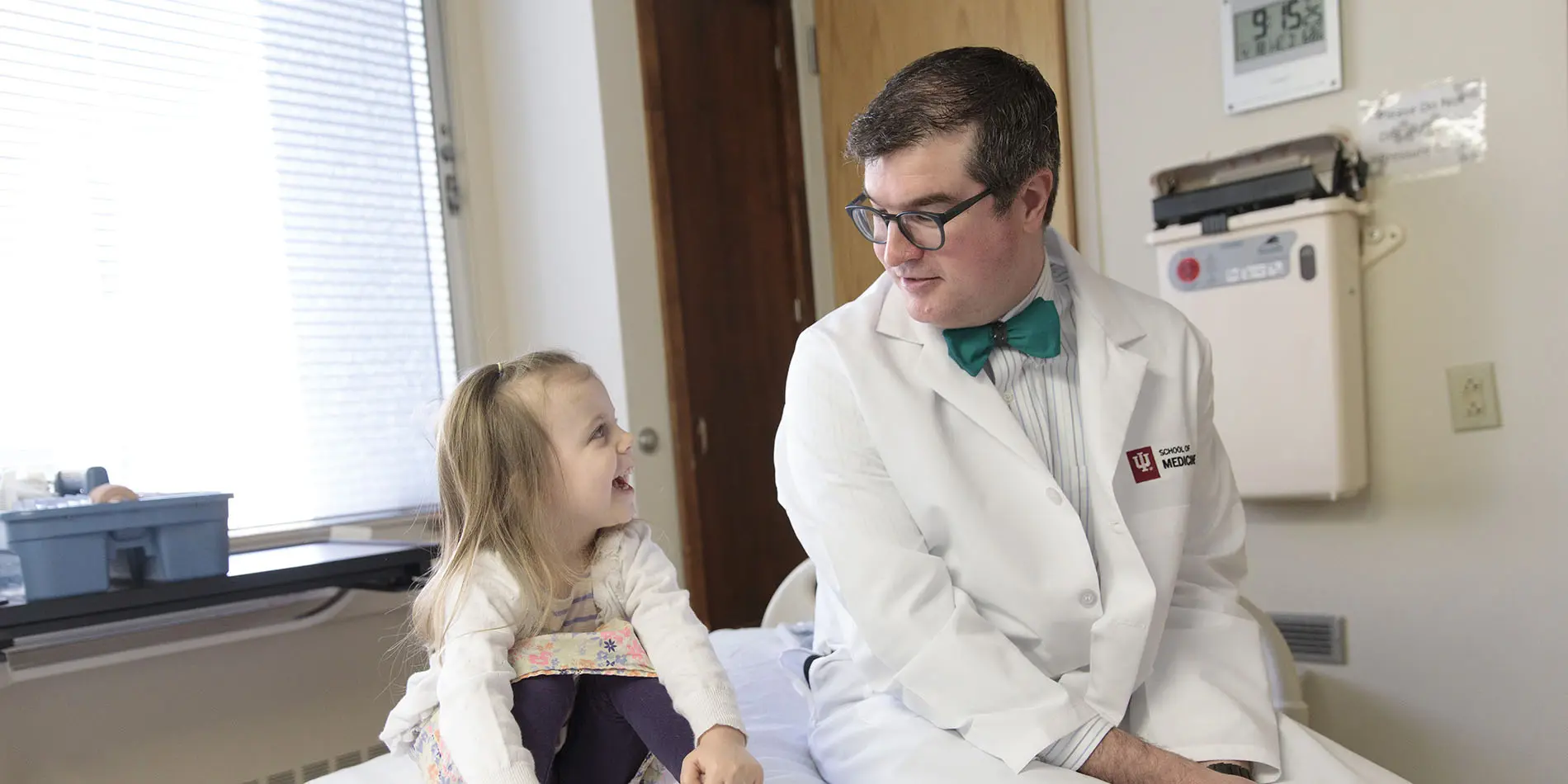A physician in a white coat, bow tie and glasses sits next to a smiling young girl in an exam room
