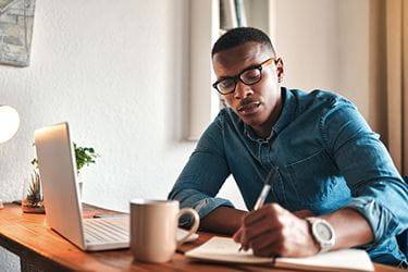 A young professional male writes notes while sitting at a desk with a laptop on the table.