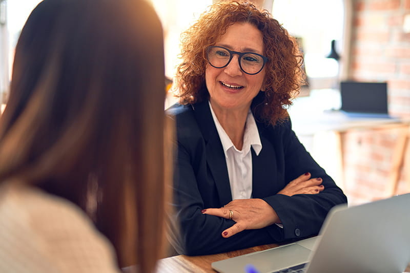 Two businesswomen talk while sitting at a desk. They appear to be in the middle of a faculty annual review.