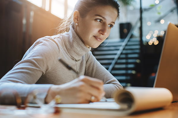 A woman takes notes on a notepad while watching a webinar on her laptop.