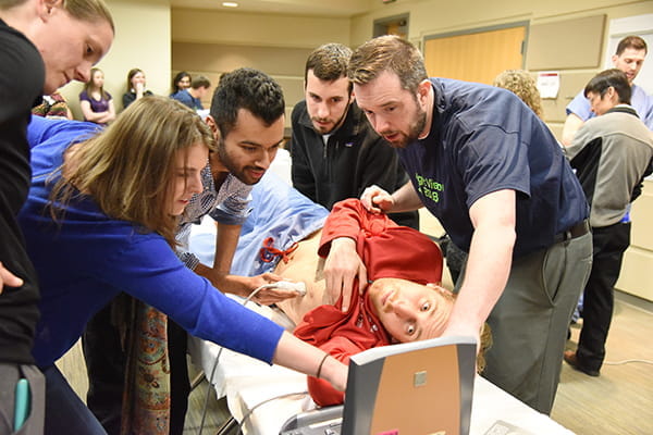 A group of students examines an ultrasound machine as one of the students performs an ultrasound on a male student-patient's heart