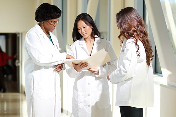 Three female physician faculty members stand in a hallway, reviewing a folder while discussing business matters.