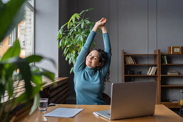 A woman sitting at a desk next to a window stretches gently, with a smile on her face. The image displays a wellness-at-work concept.