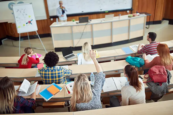 A female lecturer stands in front of a classroom while a peer reviewer (unseen) watches from the back of the room.