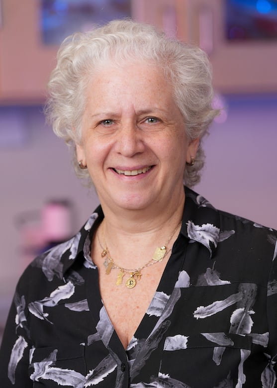 lilian plotkin wearing a black and white floral print shirt and gold necklace. She has short white hair pulled back from her face.