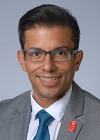 Ruben Hernandez has dark hair and glasses. He is wearing a gray suit and teal tie.
