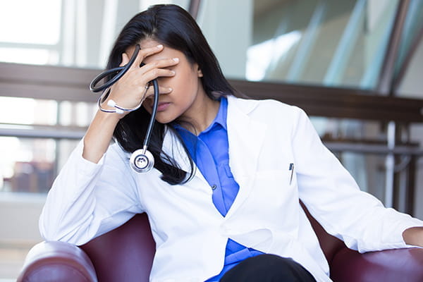 A stressed female physician holds a stethoscope and her head in her hands