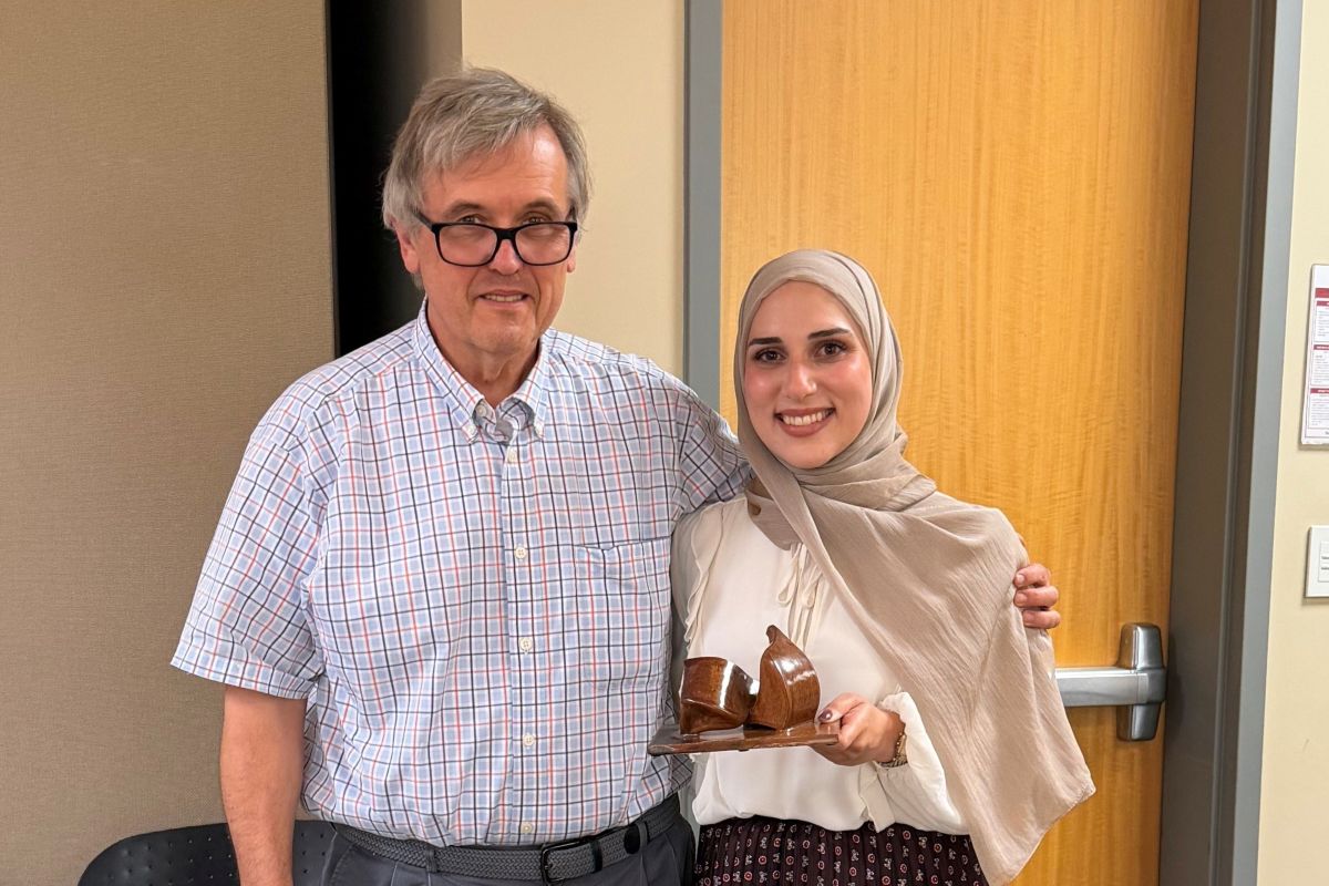 Two people pose in a classroom; one of them is holding a three dimension art piece.