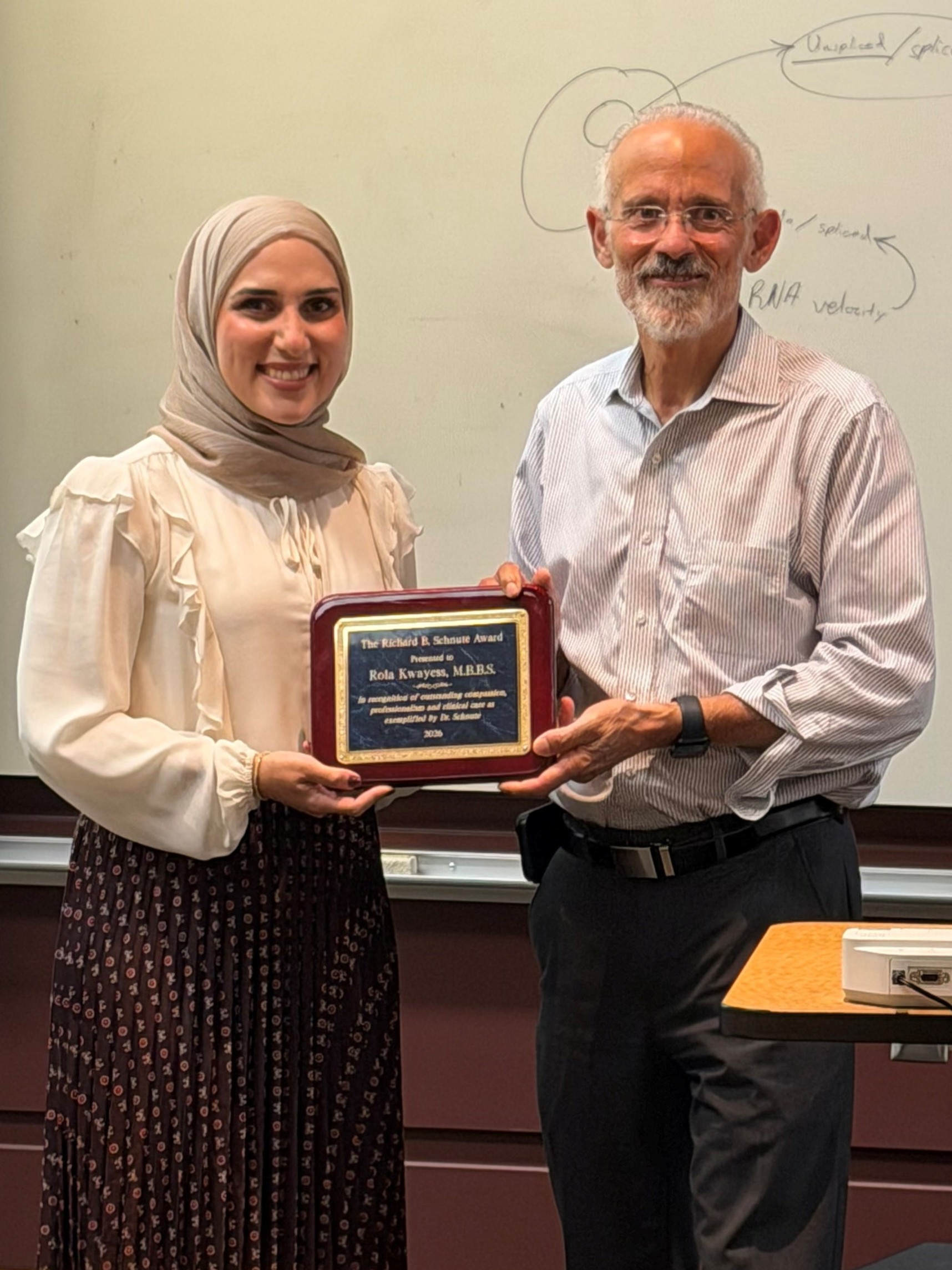 Two smiling people stand next to each other, one is holding an award plaque.