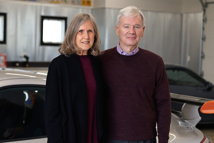 an older white woman and man stand together in front of a luxury car