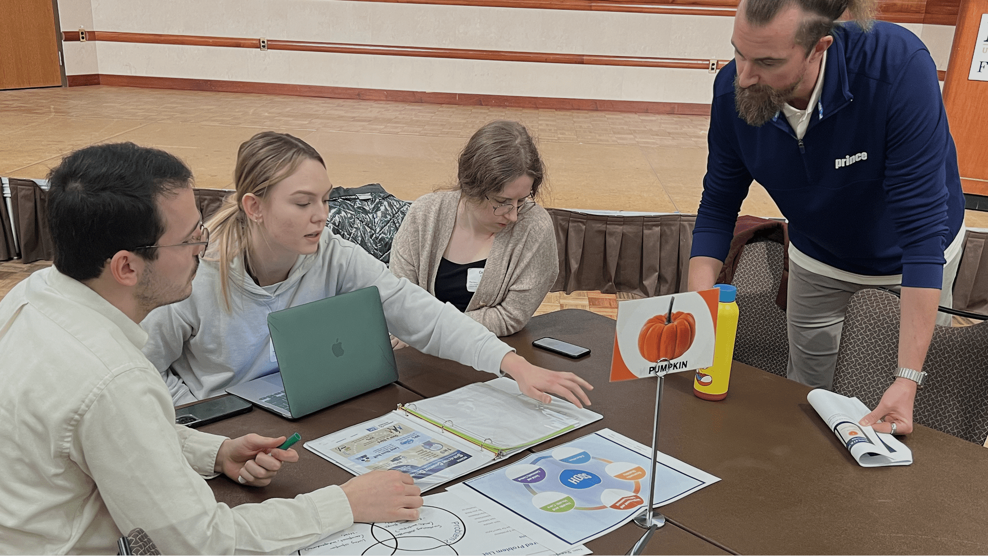 Four individuals seated around a table engaged in discussion with documents, diagrams, and a laptop, including a labeled “Pumpkin” diagram.