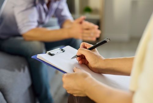 image of a person filling out paper on a clipboard in front of a man sitting on a couch