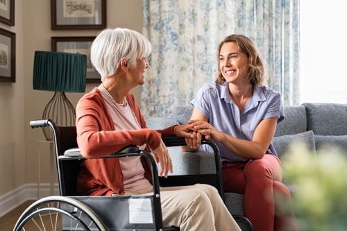 white elderly woman in wheelchair holding hands with female caregiver sitting on couch