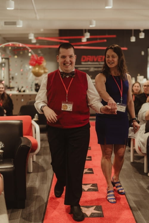 man with intellectual disability in red vest and black pants walking down a red carpet holding female caregivers hand and leading  