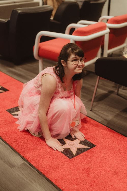 picture of a woman with a disability in a pink dress bending down touching a star with her name on it on a red carpet