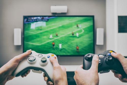 two sets of hands playing a soccer video game, one holding a white controller the other holding a black controller