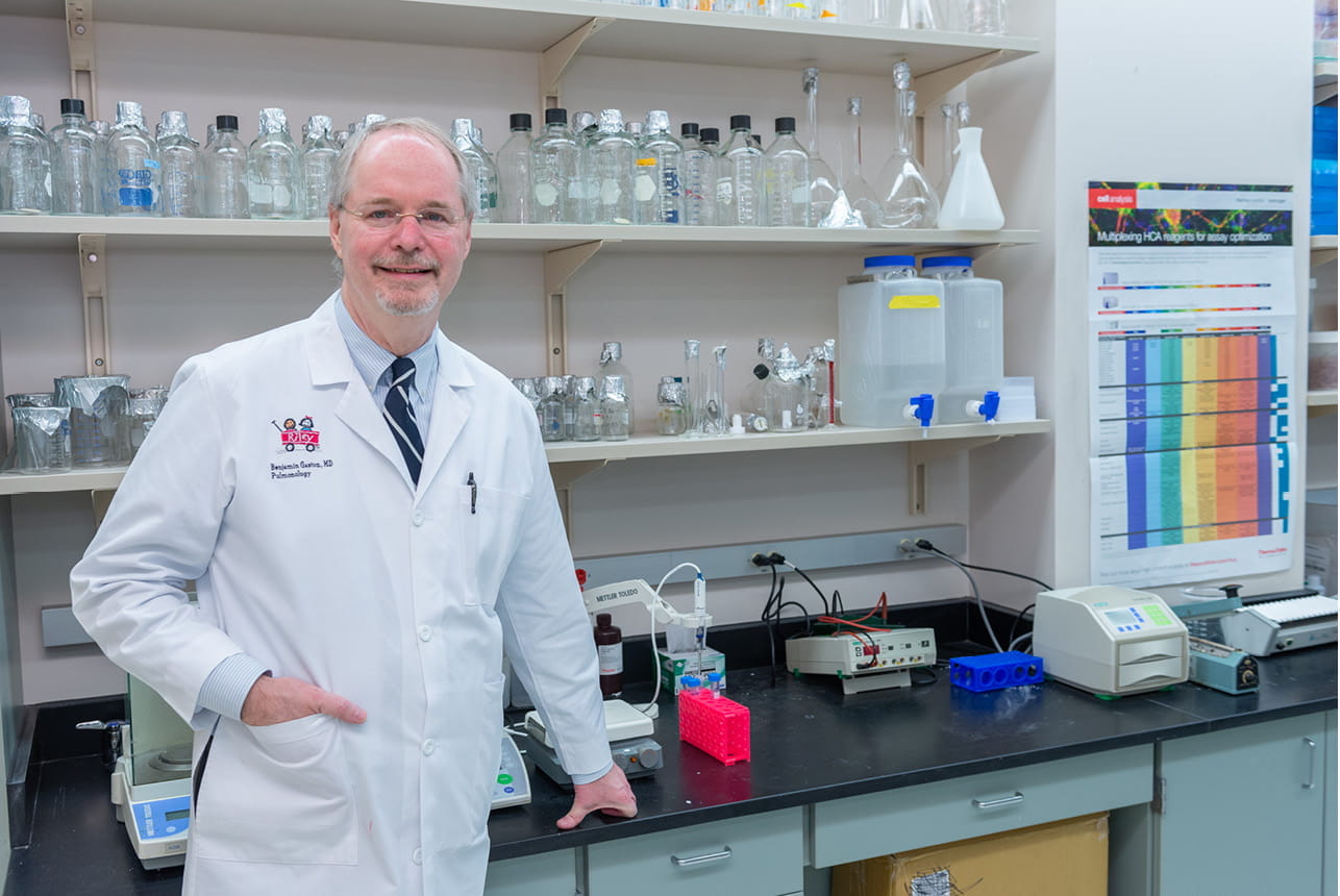 Benjamin Gaston stands in his lab wearing a white coat and glasses. He has light skin, white hair and a white beard.