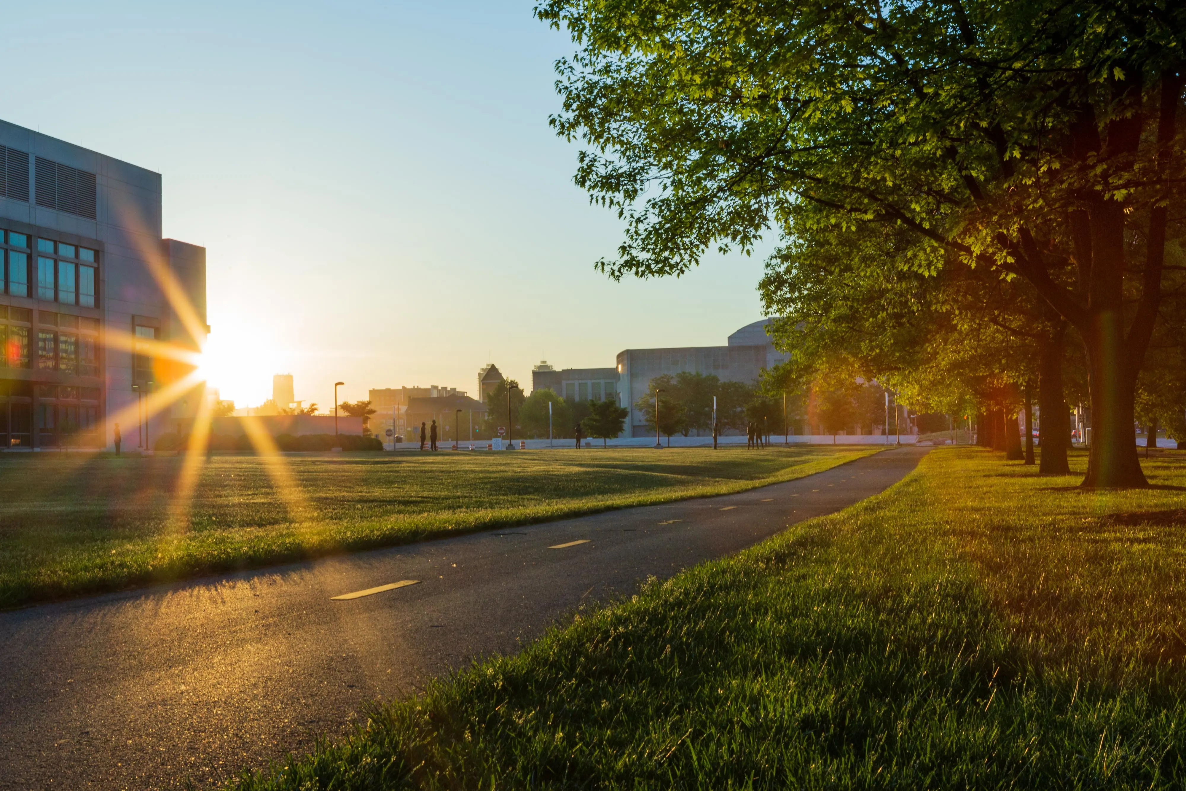 Sunrise over a tree-lined walking and biking path in downtown Indianapolis, with campus buildings and green space in view.