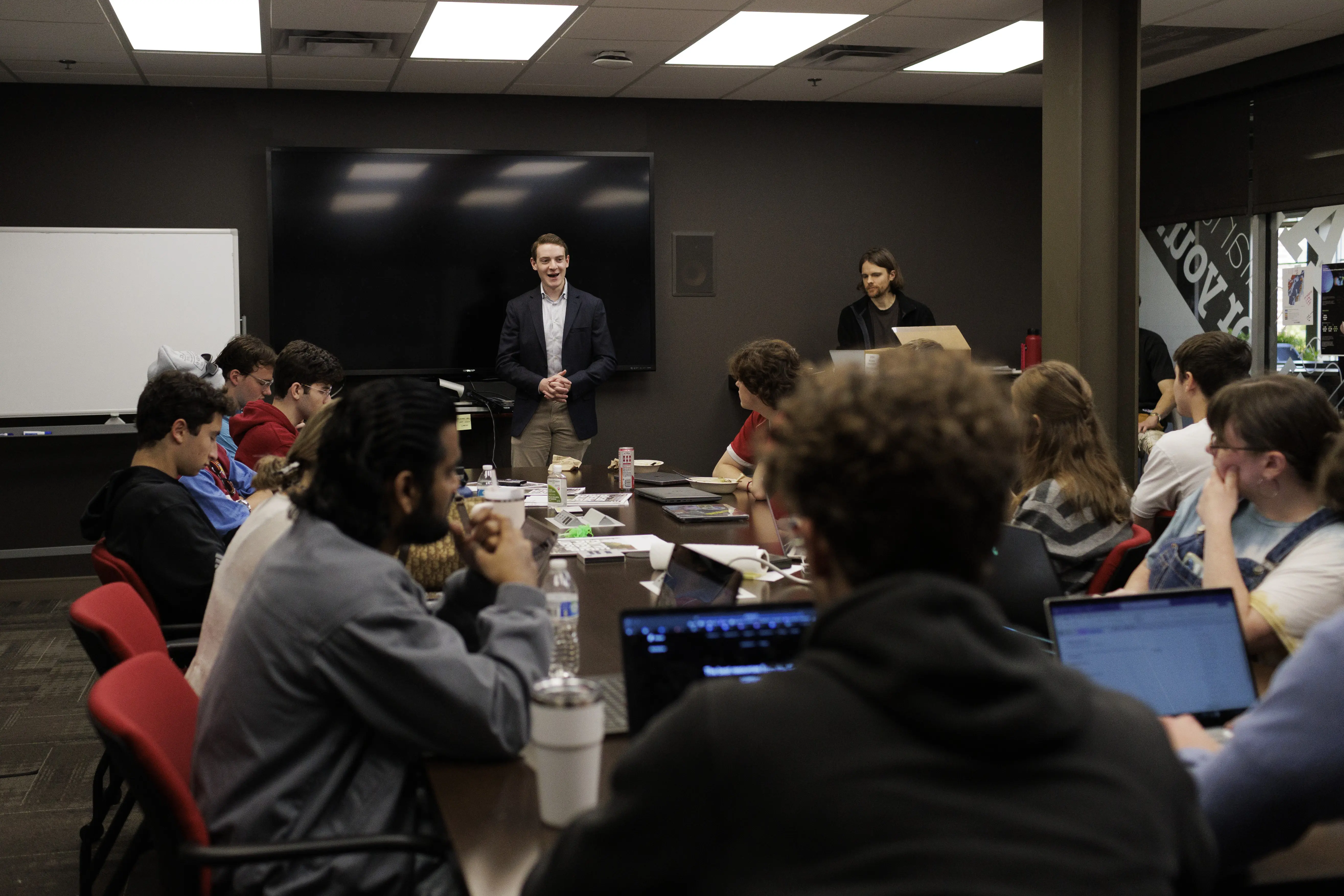 Group discussion or teaching session with participants seated around a table and a presenter at the front of the room.
