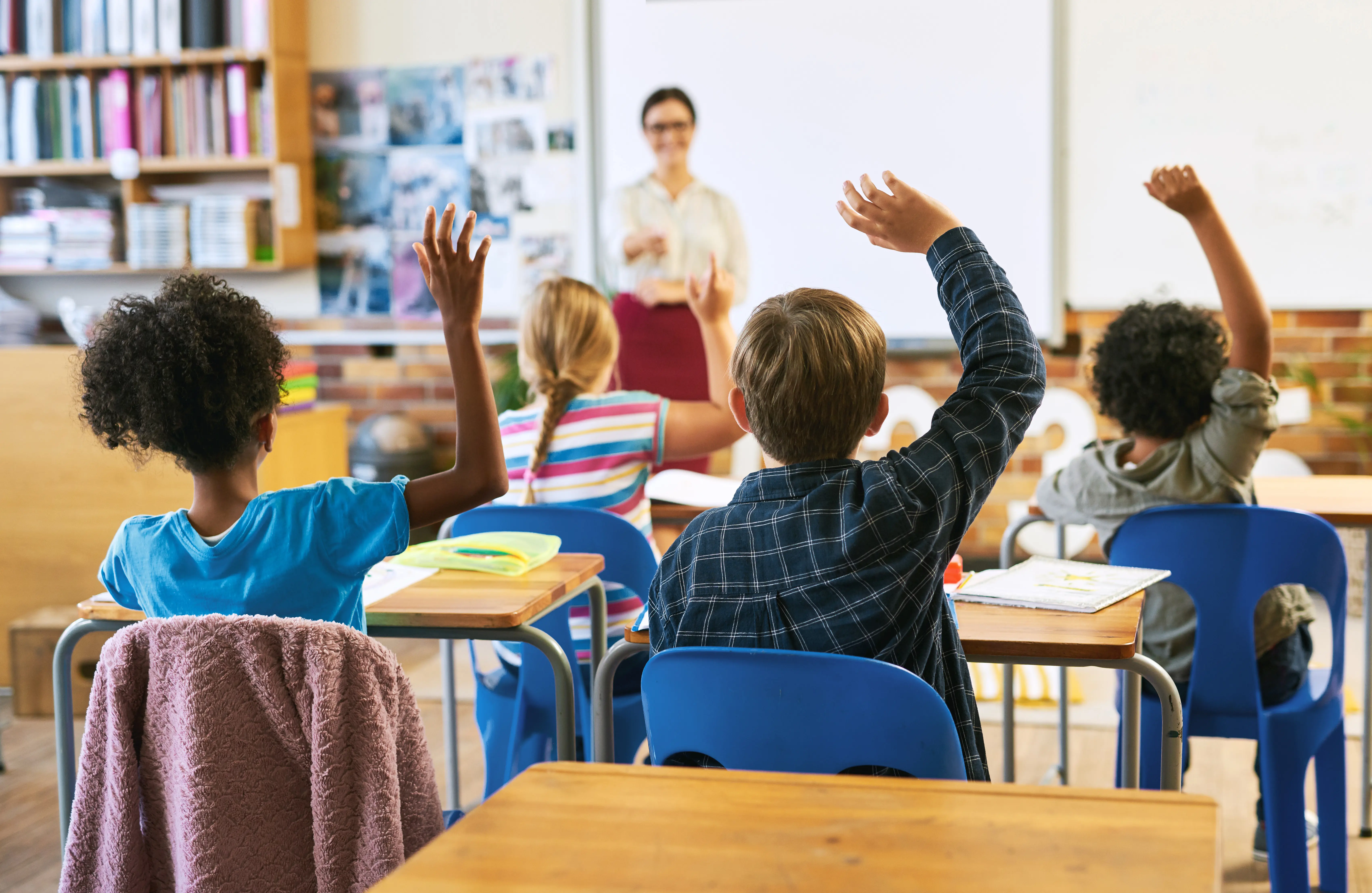 Children in a classroom raise their hands as the teacher stand in front