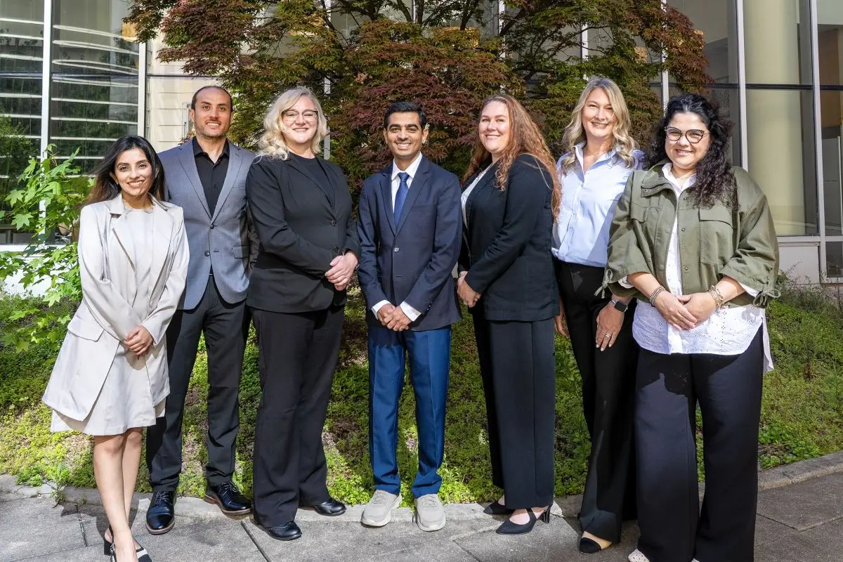 Seven smiling people pose together in front of a building and green landscaping.