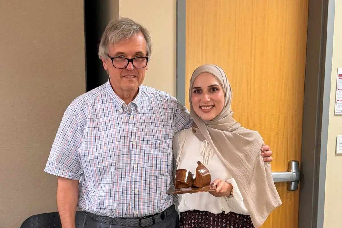 Two people pose in a classroom; one of them is holding a three dimension art piece.