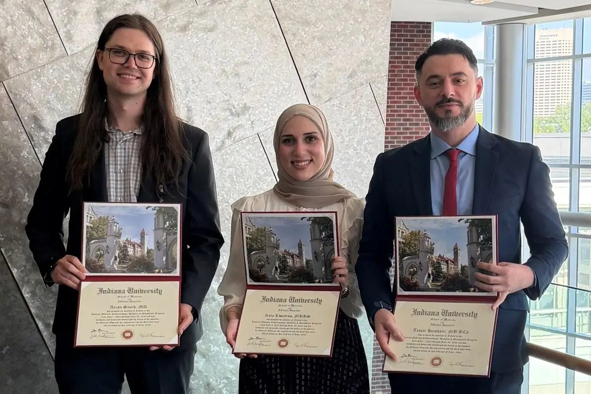 Three smiling people stand in a sun-lit hallway, holding graduation certificates.