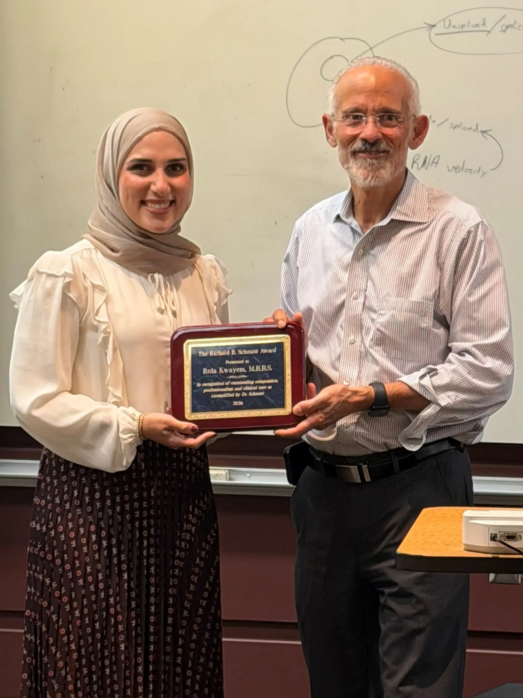 Two smiling people stand next to each other, one is holding an award plaque.