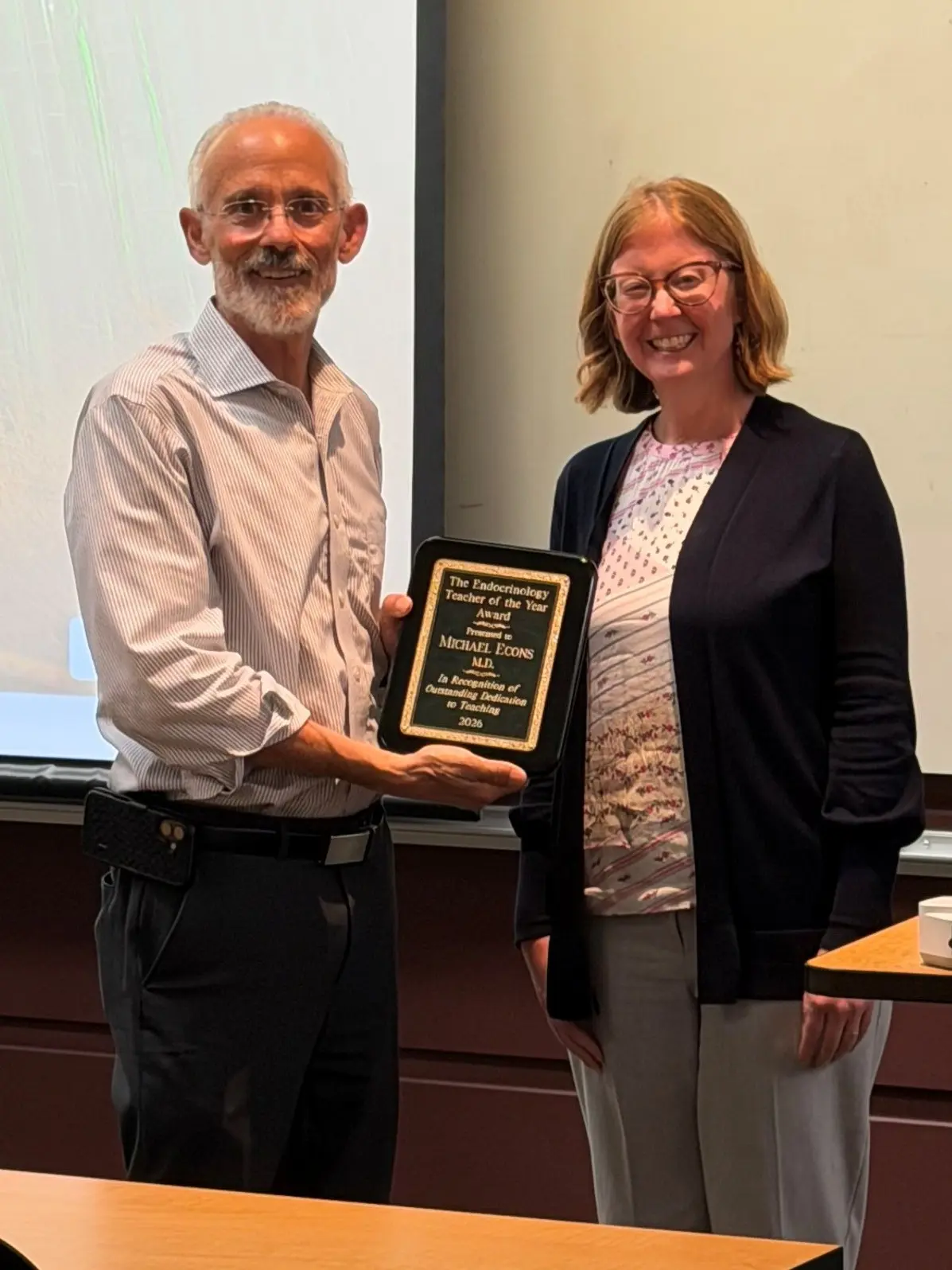 Two smiling people stand next to each other, one is holding an award plaque.