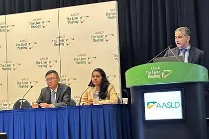 Two panelists are seated at a table with microphones while another speaker stands at a podium displaying the AASLD logo.