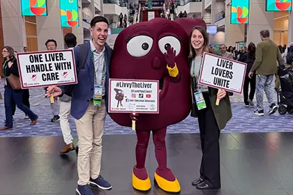 Three conference attendees pose with a person in a large liver mascot costume in a convention center atrium.