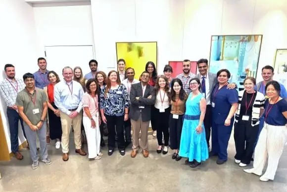 About 25 participants wearing conference name badges stand together in a bright indoor event space with artwork displayed on the walls behind them.