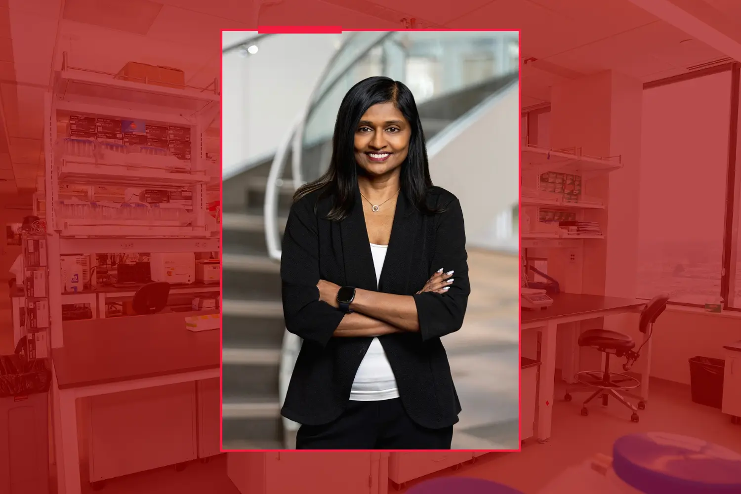 Meena Madhur, smiling with her arms crossed, wearing a black blazer and white shirt, against a red-toned background of a lab.