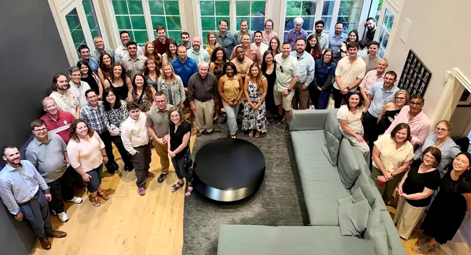 Large group of people gathered indoors in a bright room with tall windows, arranged around a dark circular table and a gray sectional sofa, posing for a group photo taken from an elevated angle.