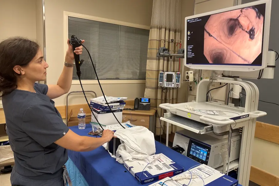 Healthcare professional holding an endoscope connected to a cart with medical equipment, while a monitor displays an internal view from the scope.
