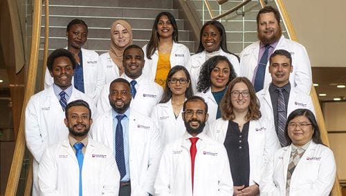a group of 15 diverse residents wearing white coats stand together on a staircase