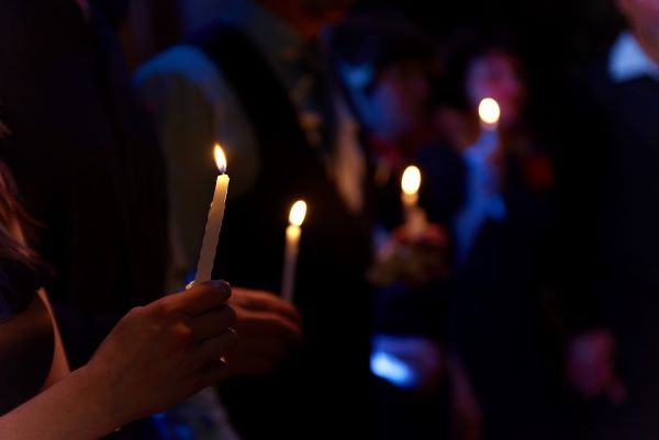 Hands holding lit candles in the dark, against a background of blurred silhouettes with candles.