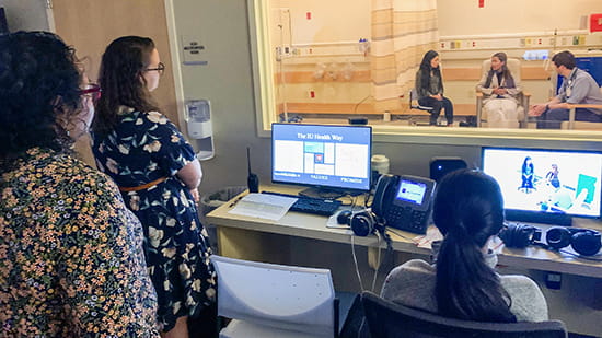 Three women watch a simulation from behind a women. In the room are a male physician and two young women.