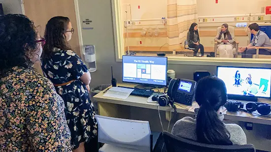 Three women watch a simulation from behind a women. In the room are a male physician and two young women. 