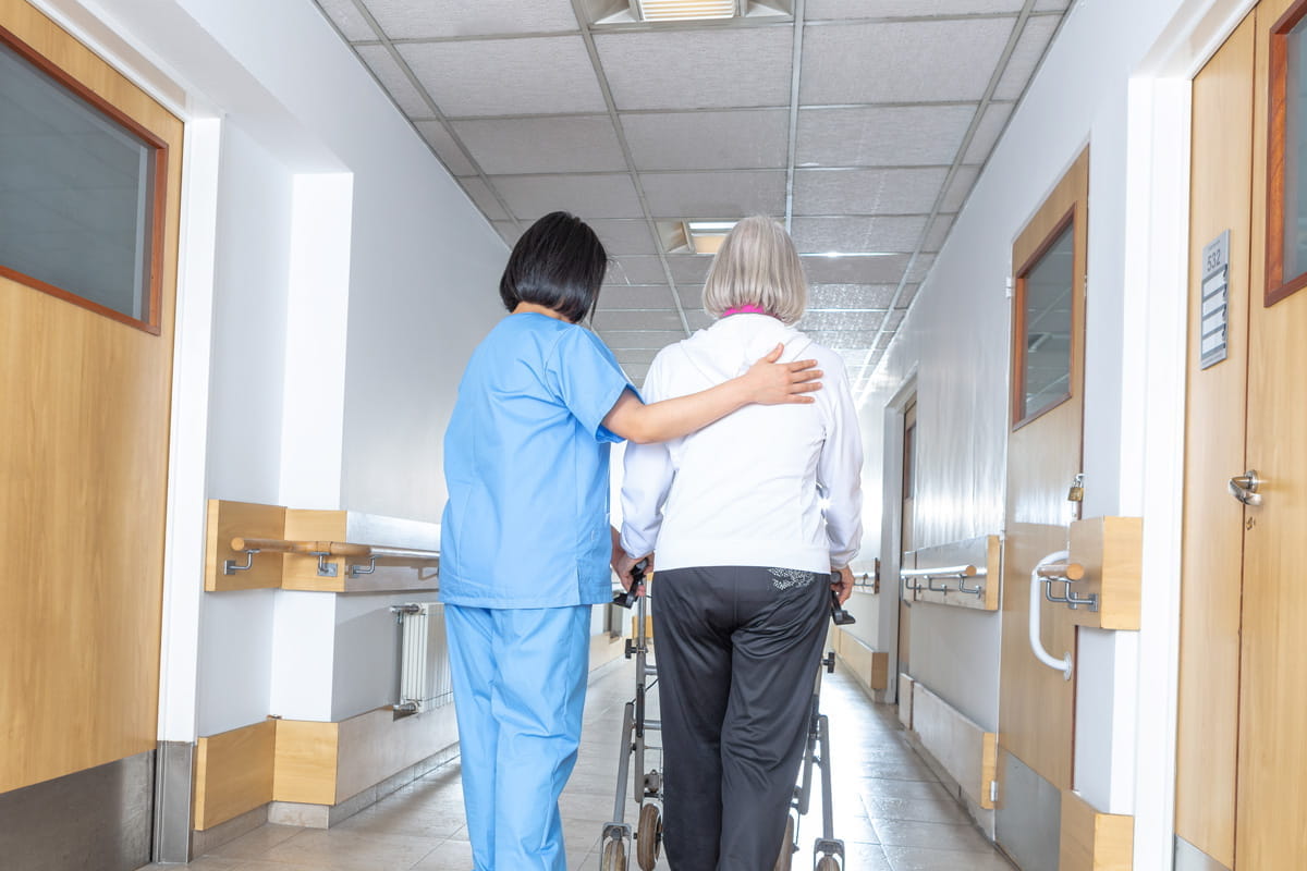 a provider helps an elderly patient walk down the hallway using a walker