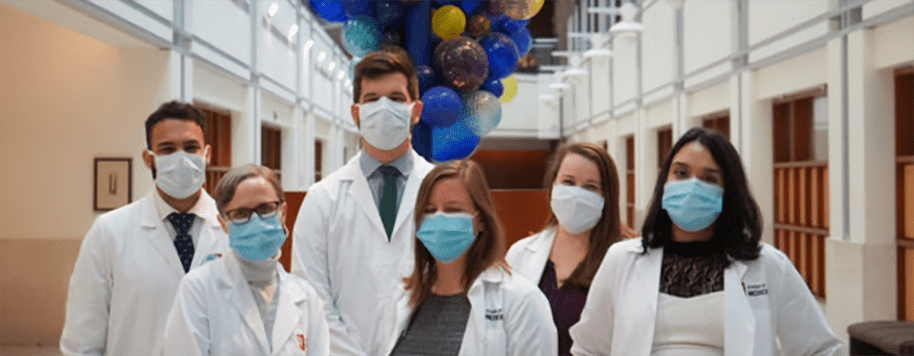 6 Former INFD Fellows. The background is the Van Nuys Medical Science Building interior with a large sculpture in the background. Each fellow is wearing a surgical mask and is wearing a lab coat.
