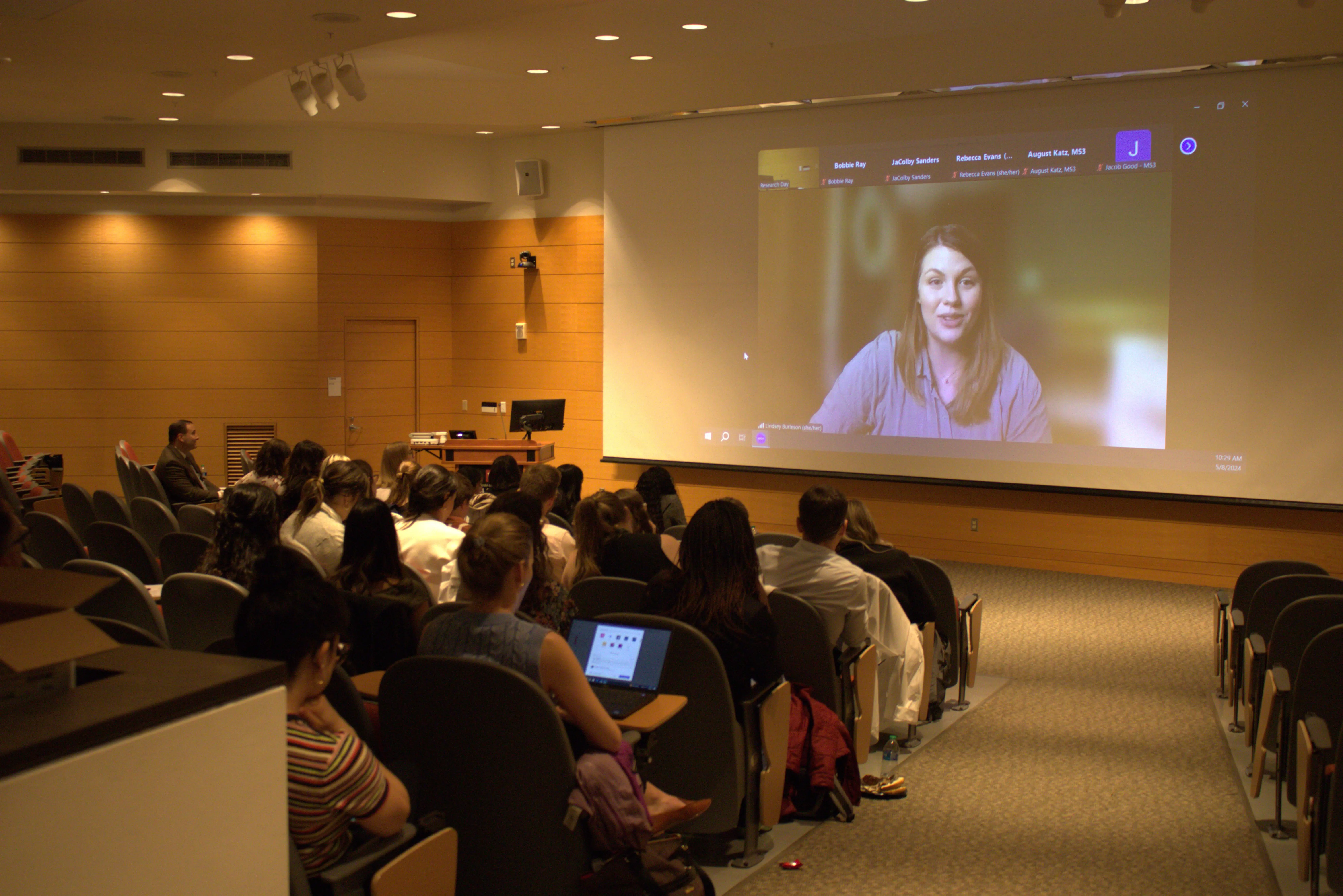 Dr. Lindsey Burleson smiles on the projector while her audience in the auditorium watches