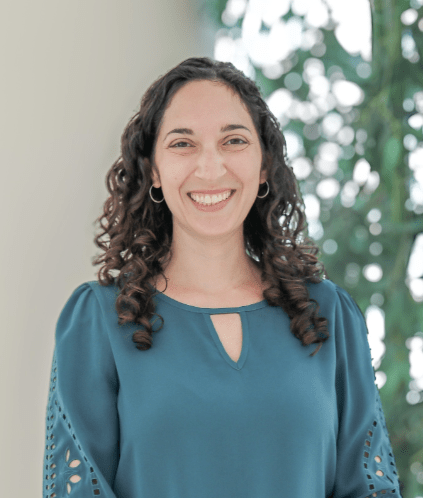 Cara Berg smiles for her headshot in front of a concrete pillar and bamboo.