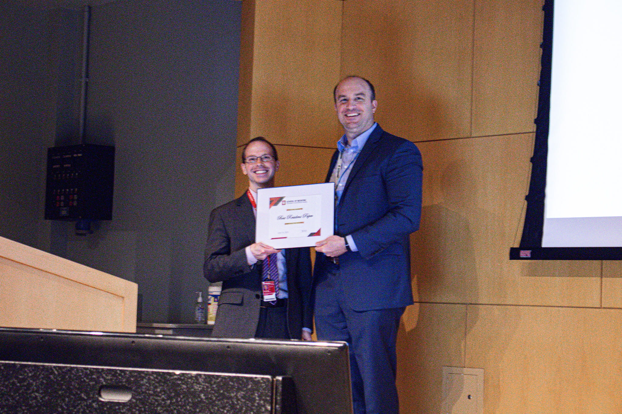 Julian Dilley stands with Luke Lopas on a stage behind a podium. Lopas presents Dilley with a paper recognizing his research excellence. Both men hold a side of the paper.